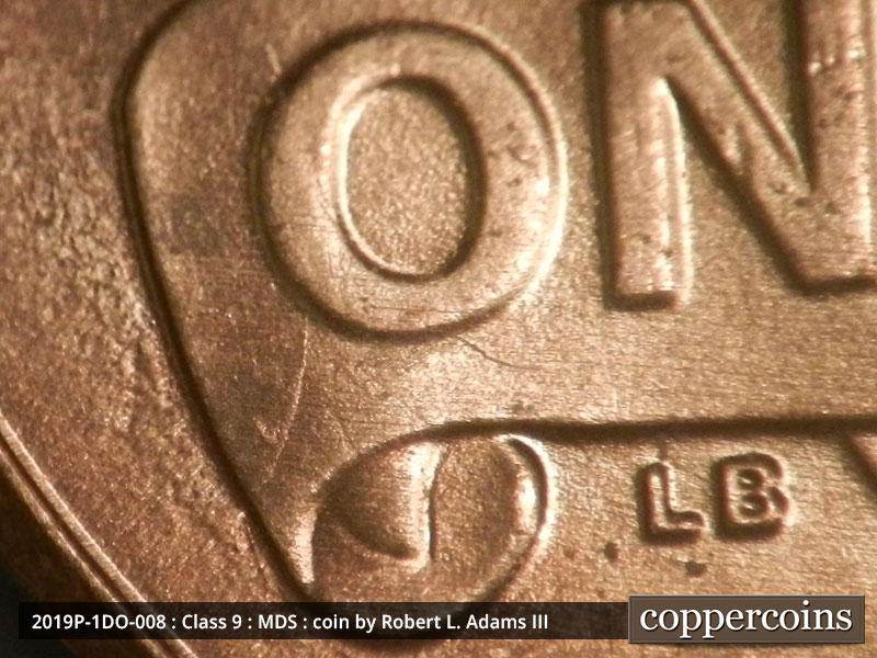 Die-stage B marker on 2019P•1DO•008 showing They died chip forming on the left side of the banner through the O.